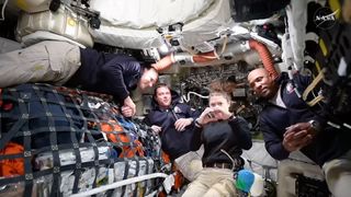 four people in black sweatshirts inside a cramped spacecraft cabin packed with cargo held down by netting and tubes and wires