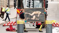 Two firefighters wearing gloves carry a large framed painting of two nude men in front of a fire truck