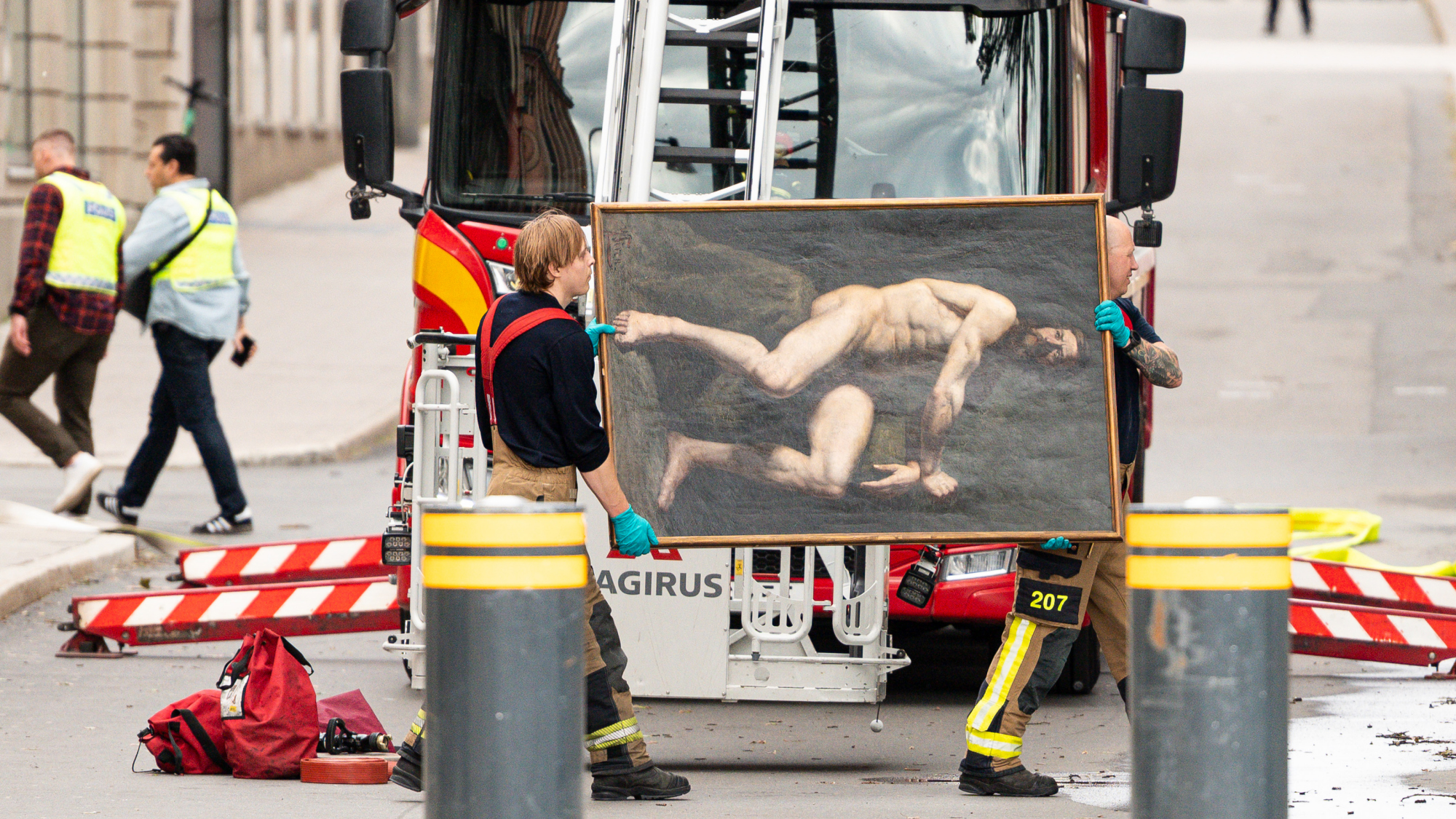 Two firefighters wearing gloves carry a large framed painting of two nude men in front of a fire truck