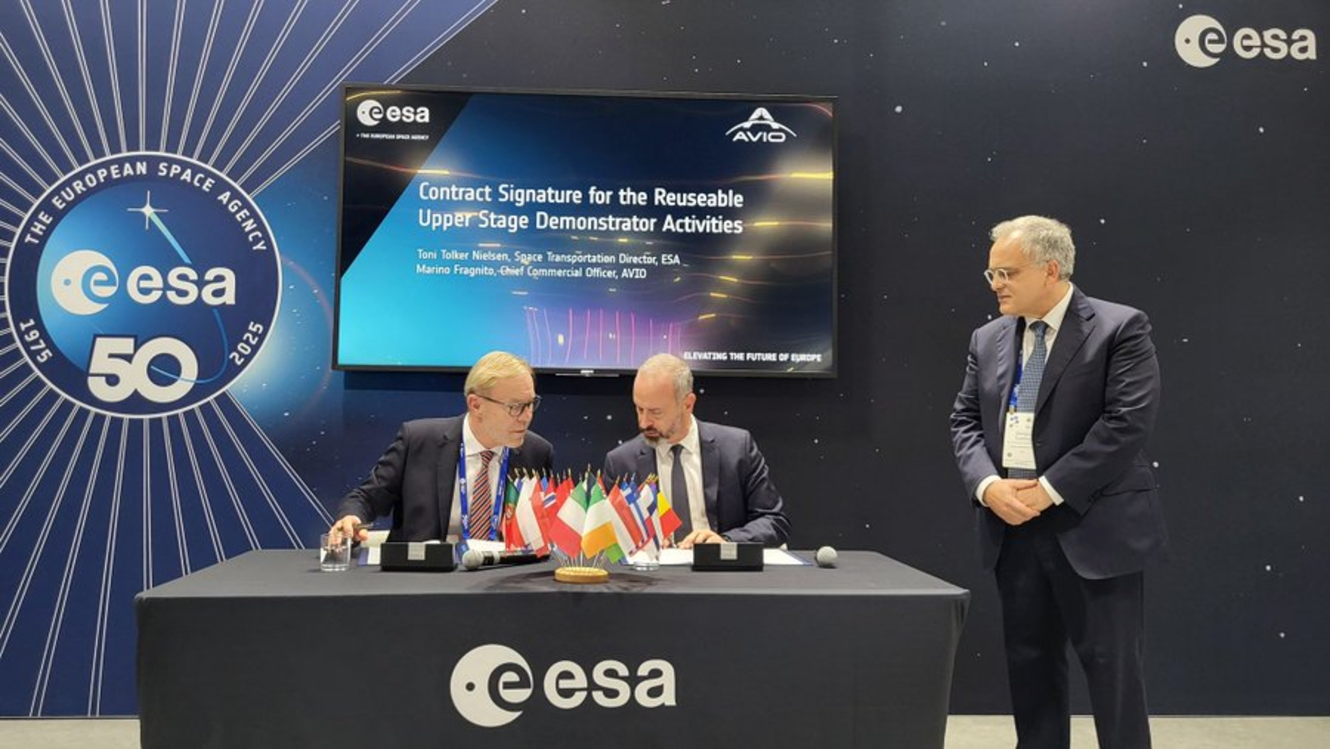 Two men sit behind a covered table with an ESA sign on it talking to each other.