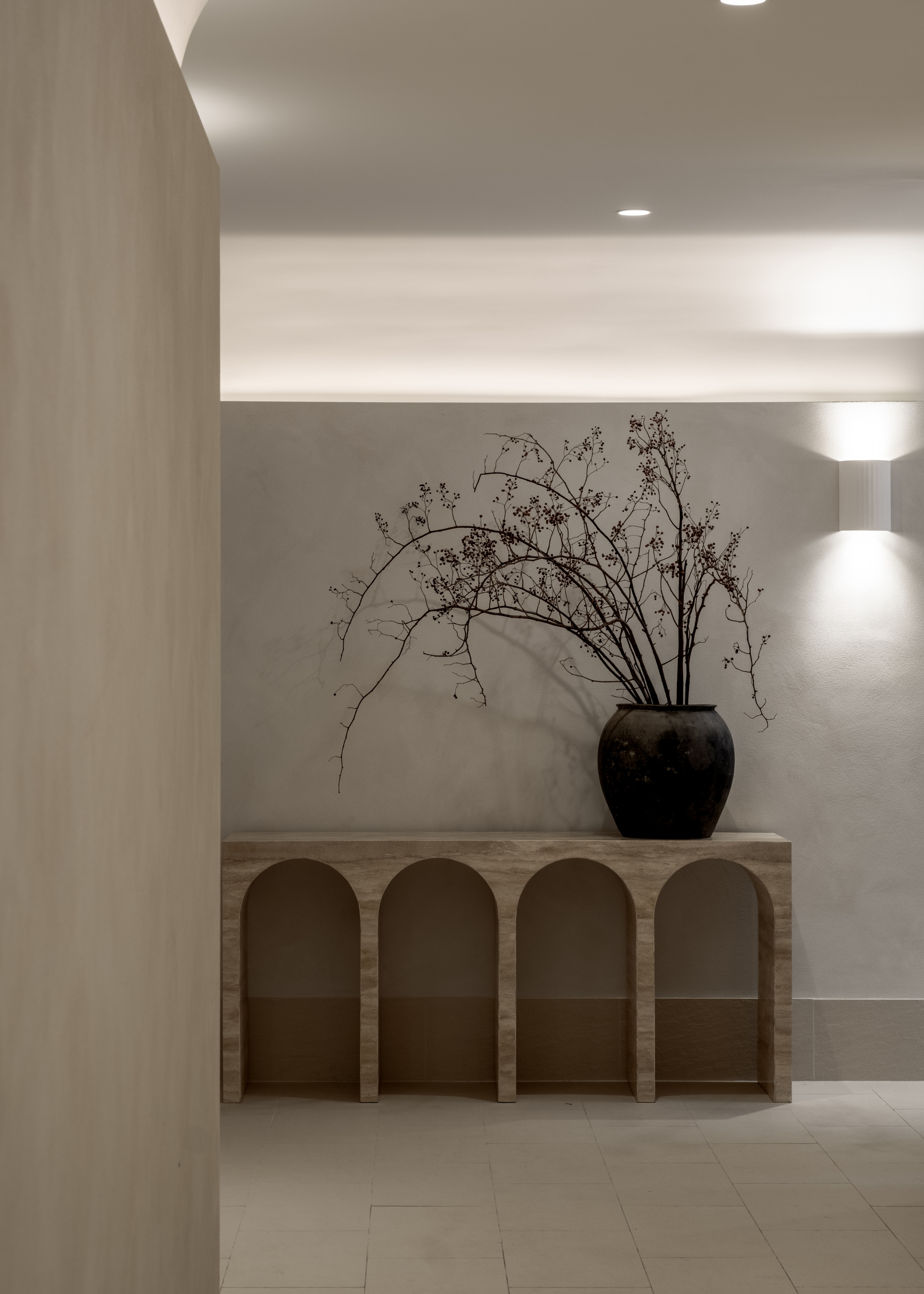 A calming entryway with an arched side table featuring a large vase of branches by a white wall sconce