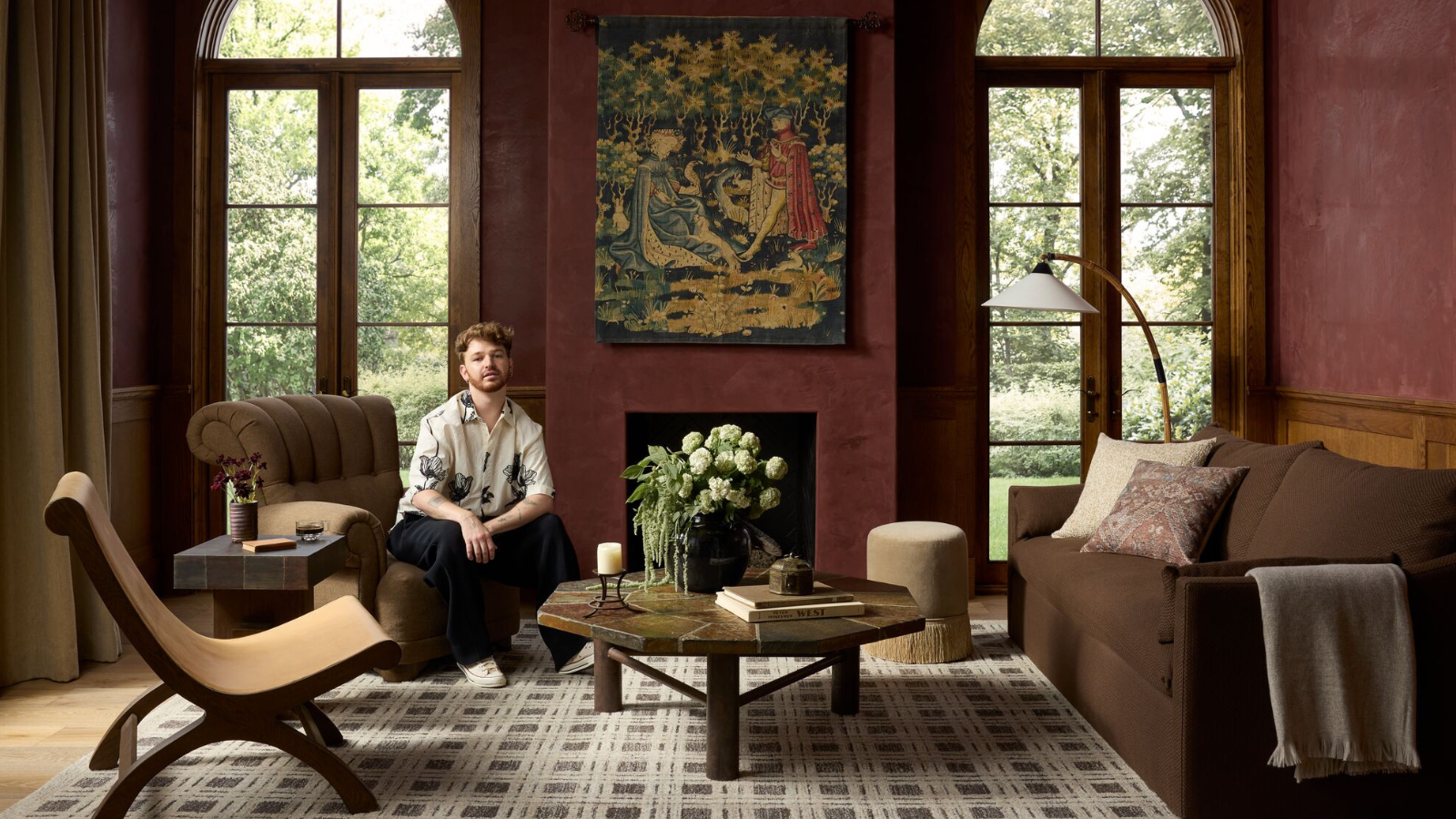 a moody, medieval-inspired living room with a brown and gray checker printed rug, a chocolate brown sofa, two accent chairs, red painted walls, a vintage-inspired tapestry, and a man sitting on one of the chairs
