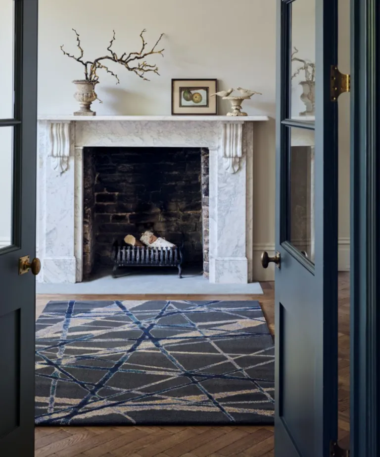 A large white fireplace with wooden floors, a dark grey patterned rug in front of the hearth