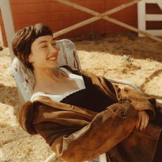 Image of Sydney Chandler for her October In Focus story sitting in an old rusty chair wearing a look by Miu Miu. 