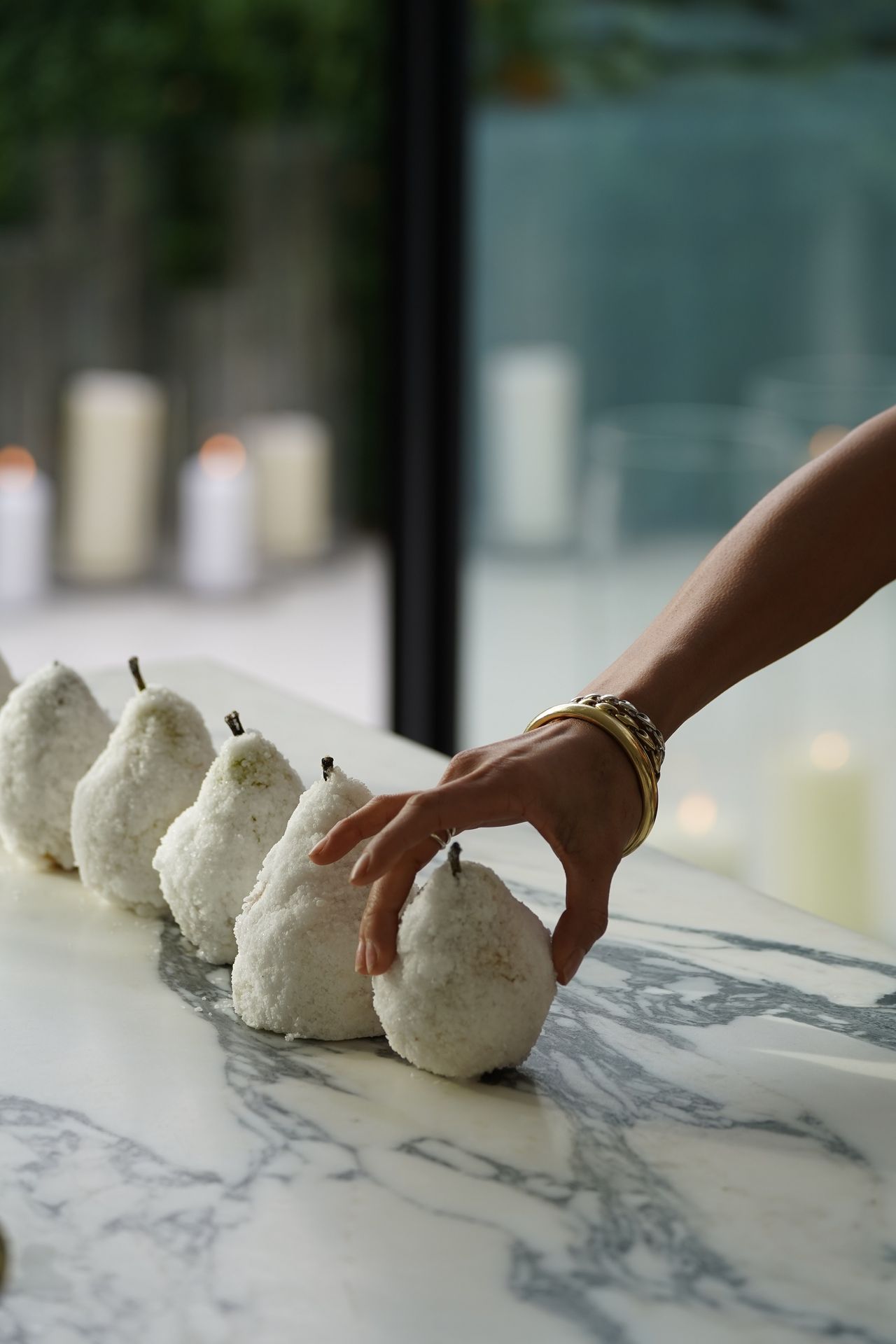 A hand is seen readjusting a series of salt-crusted pears lying on a marble kitchen counter.