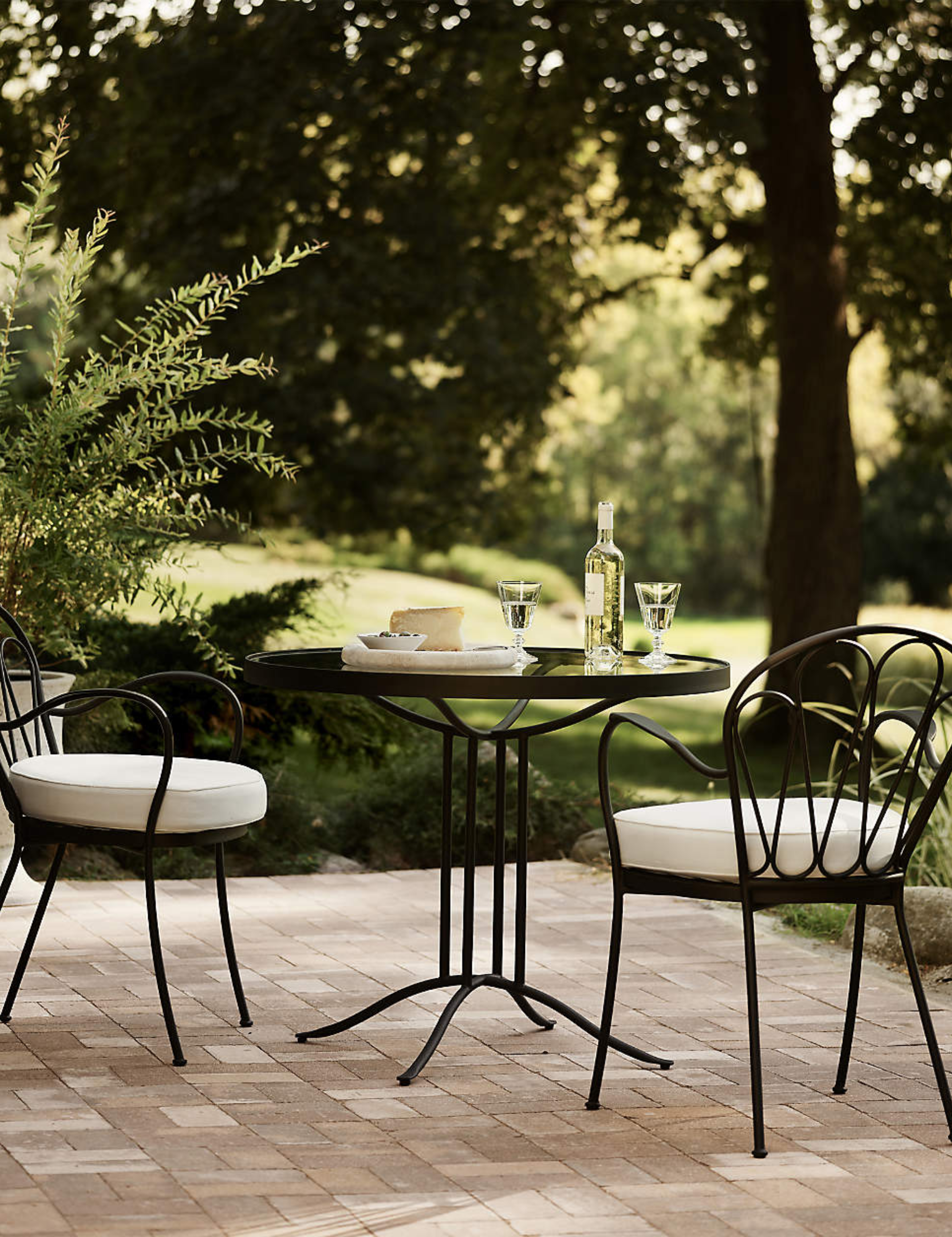 Whimsical wrought iron bistro table set topped with white wine and stemware, set atop of a brick patio overlooking a lush backyard