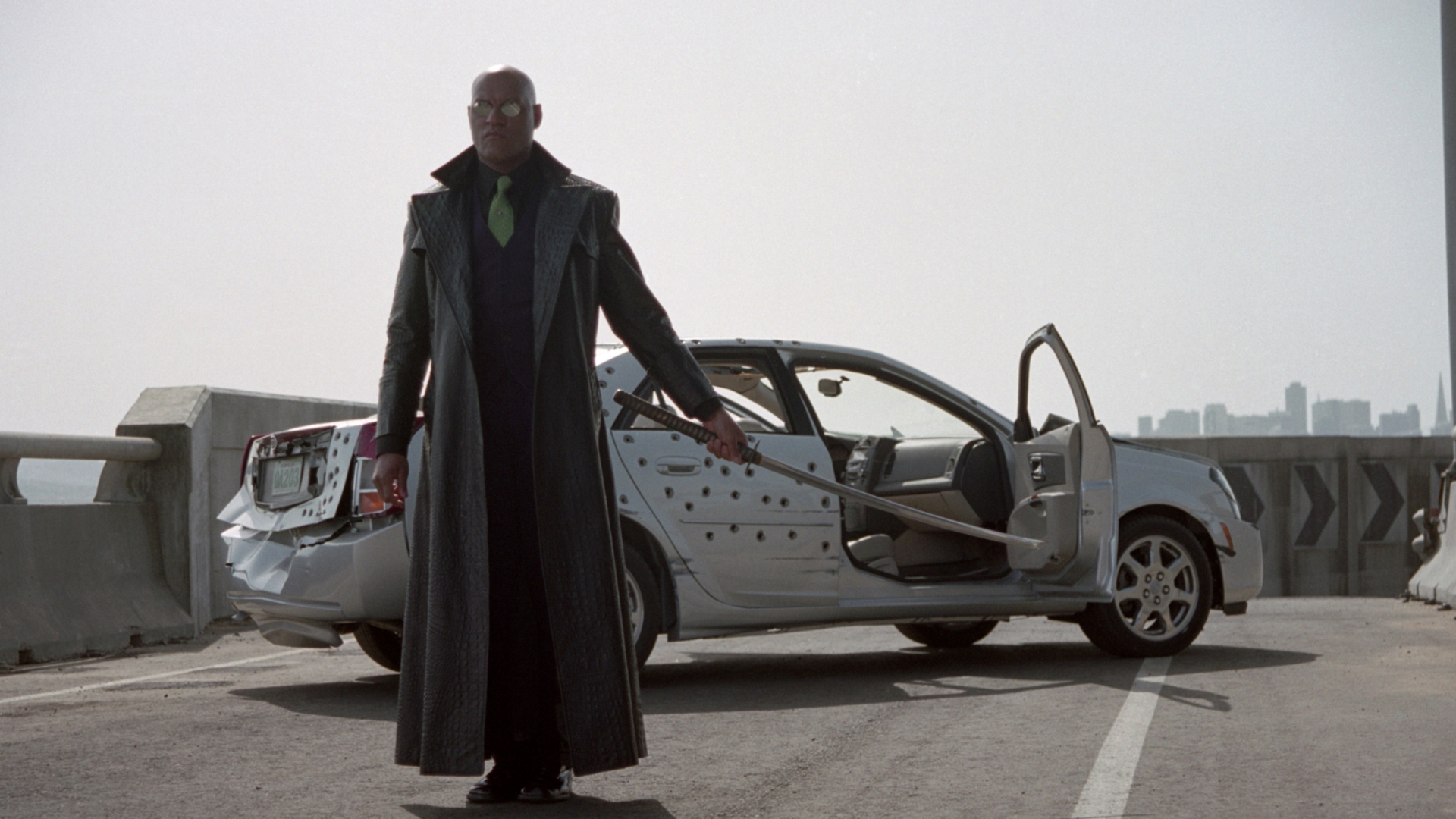A still from The Matrix Reloaded movie.  Here we see Morpheus, a man wearing a long black trench coat, green tie and reflective sunglasses, set out of a white, bullet-ridden car as he holds out a samurai sword.