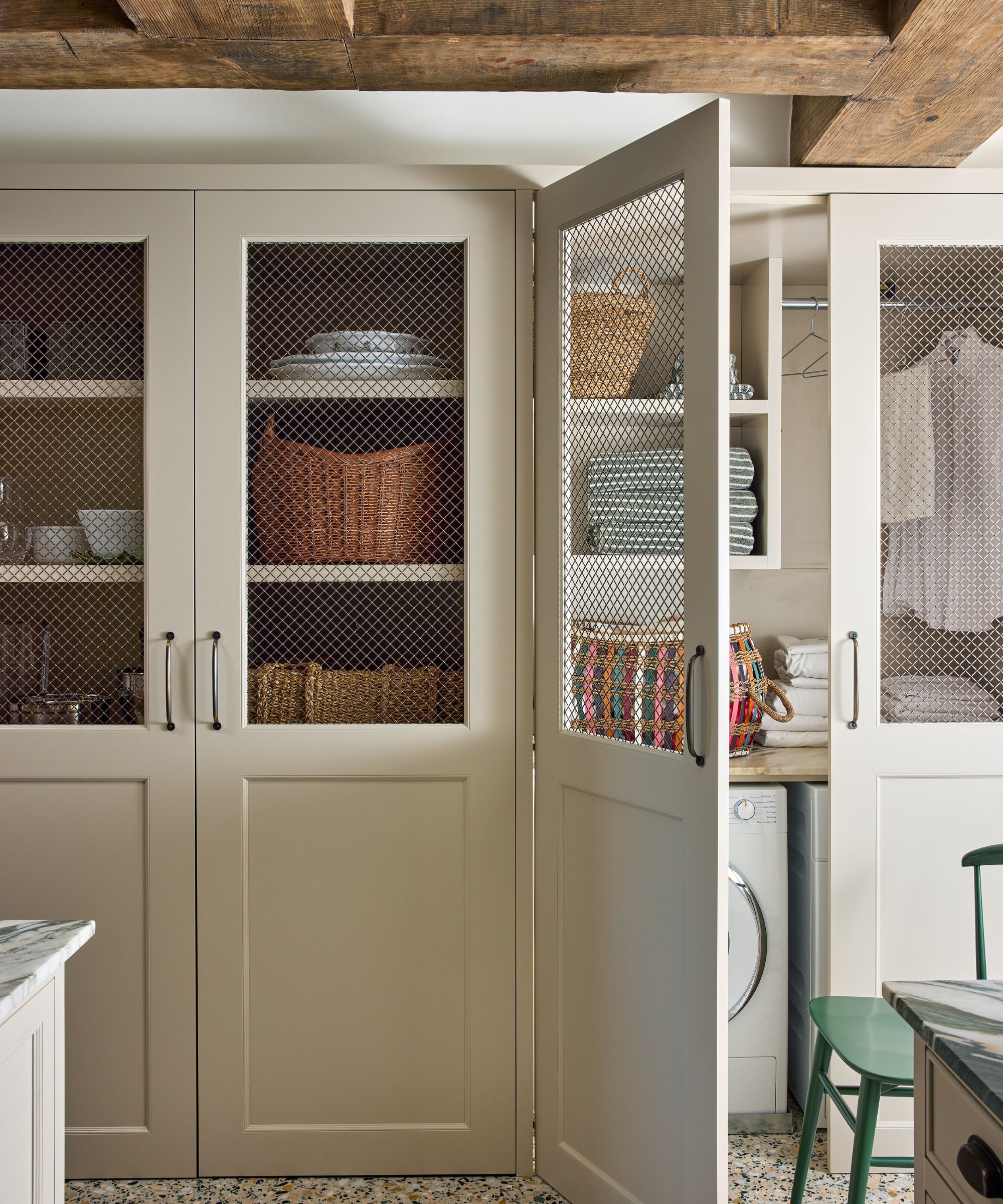 A utility space in a kitchen with full-height neutral cabinets with a mesh screen on the upper portion. Wood ceiling beams above, and a washer in the cupboard with shelves for storage.