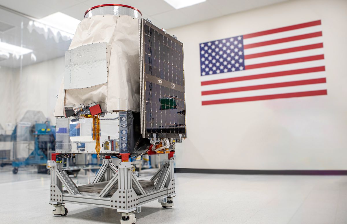 A covered metal probe is placed on a cart in a clean room and an American flag hangs on the wall behind them