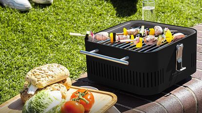 Everdure Cube BBQ by Heston Blumenthal in black on brick wall cooking skewers with rolls, tomatoes and lettuce on chopping board next to it