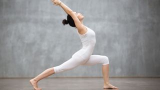 Woman in white leotard performing a high lunge during yoga routine against grey backdrop