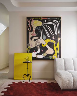 colorful living room with yellow artwork leaning against the wall, a bold artwork on the wall, the edge of a boucle sofa, and a pink and red rug with a scallop border