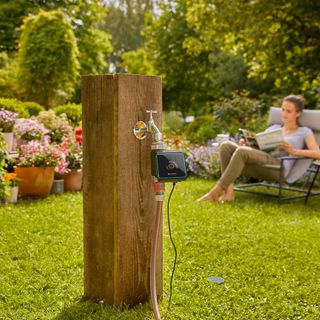wooden post in the middle of garden with outdoor tap and smart irrigation system attached to it and in the background woman relaxing on chair in garden