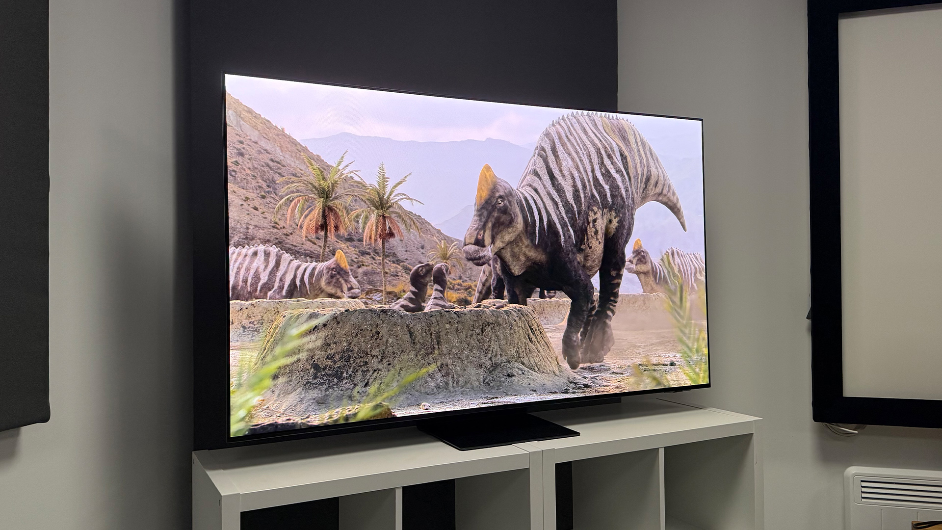 Samsung S95F OLED TV slight angle on white wooden furniture, on screen is dinosaur and baby dinosaurs