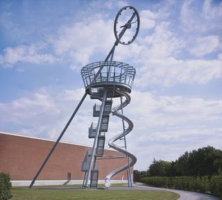 A viewing tower, with twirly slide and steps made out of steel with a transparent roof installed outside the building with trees in the background