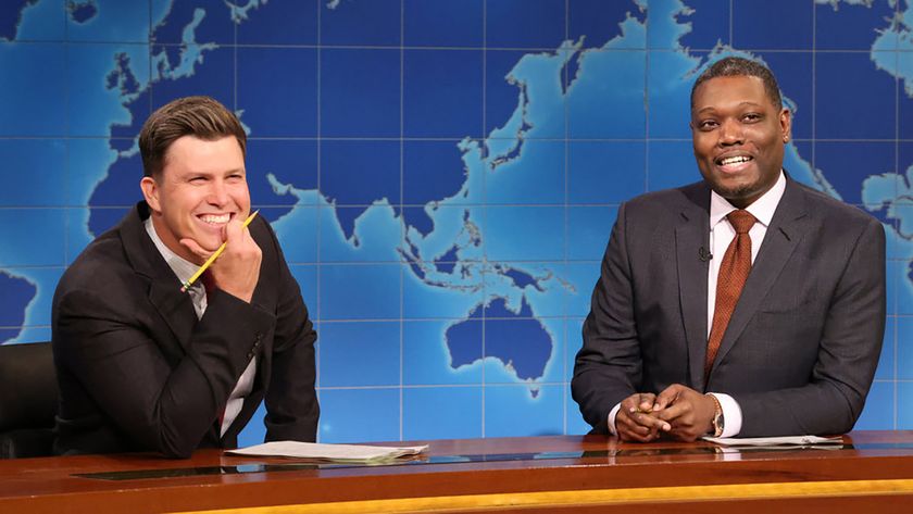 Colin Jost laughing at something Michael Che says while sitting at the Weekend Update desk on SNL.