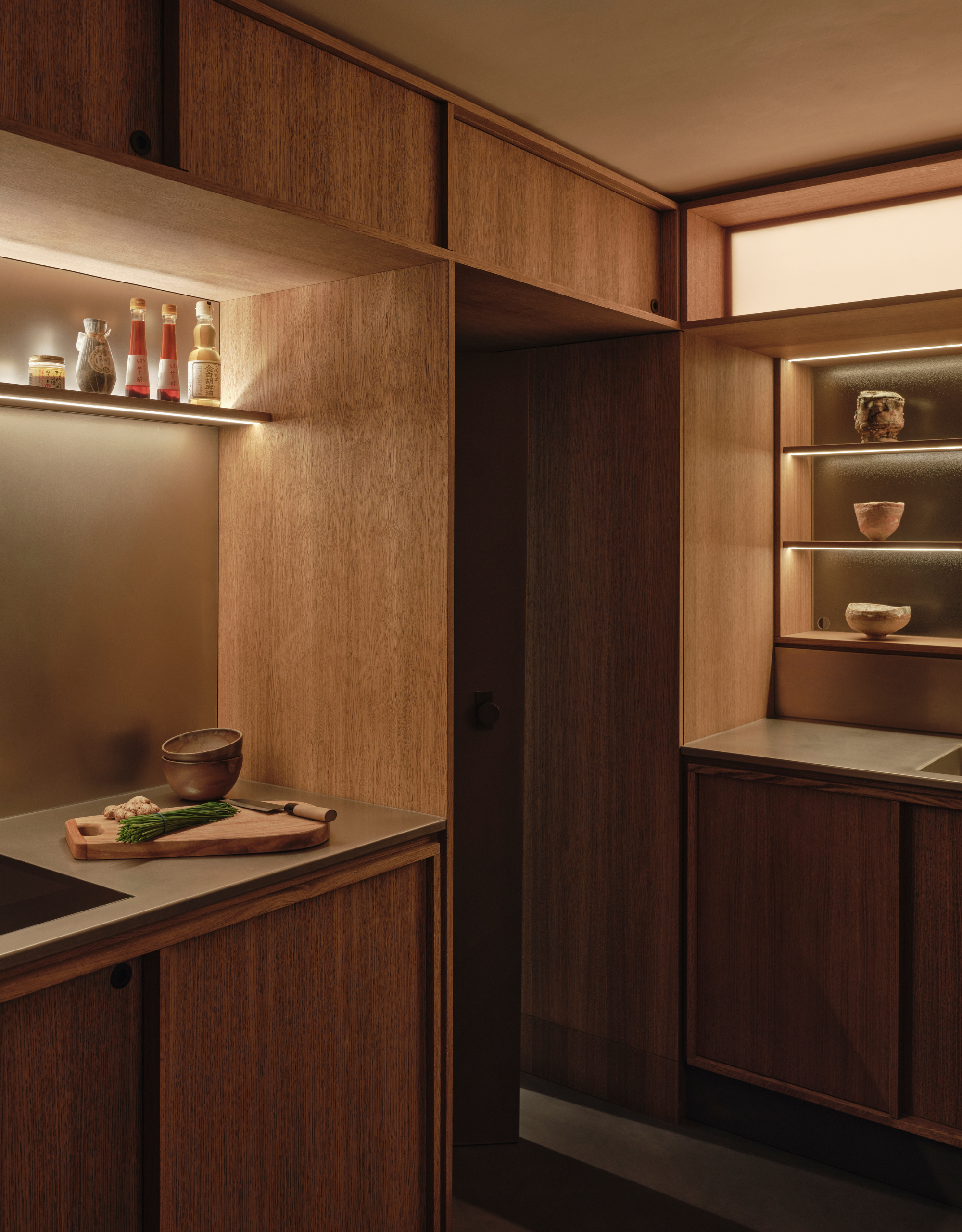 A wooden kitchen in a Japanese home with a display case of ceramics, a cutting board, and backlighting