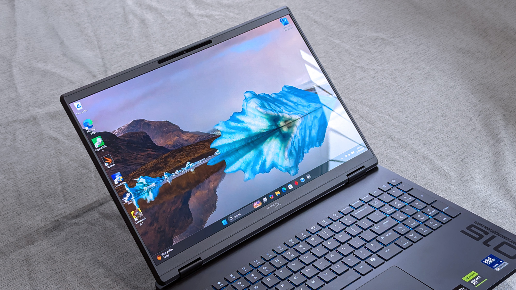 A HyperX OMEN 15 laptop displaying a Windows desktop with a scenic iceberg wallpaper, blue backlit keyboard, numeric keypad, and stickers indicating Intel Core and NVIDIA GeForce hardware.