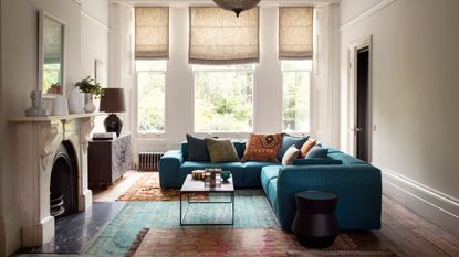 Bright living room with blue sofa, rugs and white marble mantelpiece. Behind are large windows with blinds.