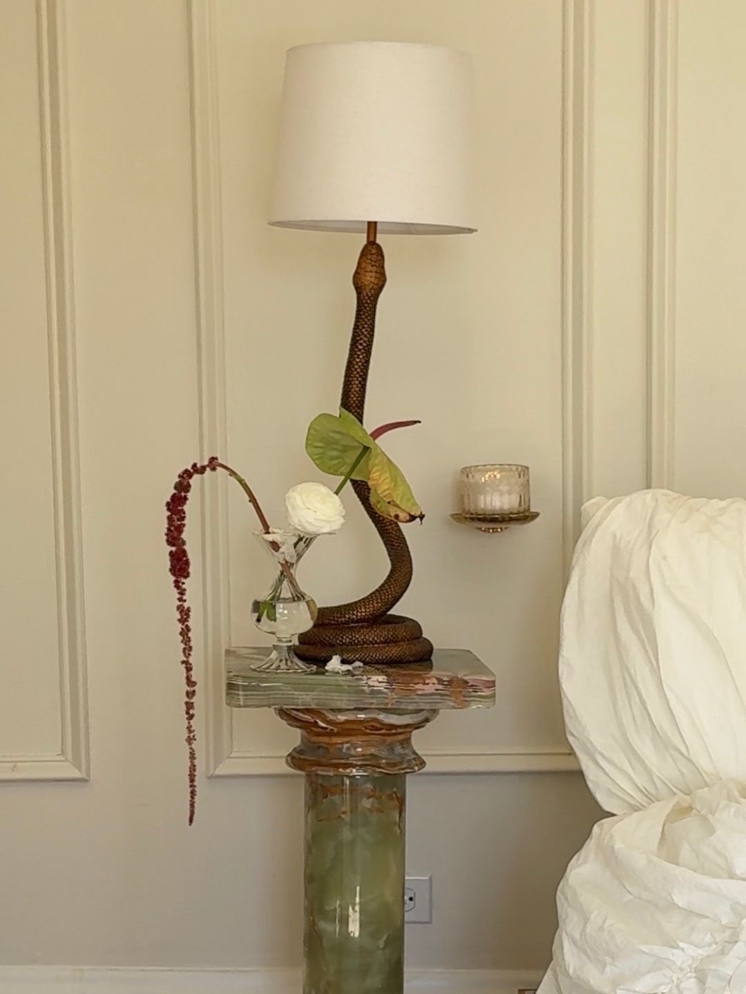 Image of a white wall with molding and a green marble bedside table that has a brass snake lamp on it.