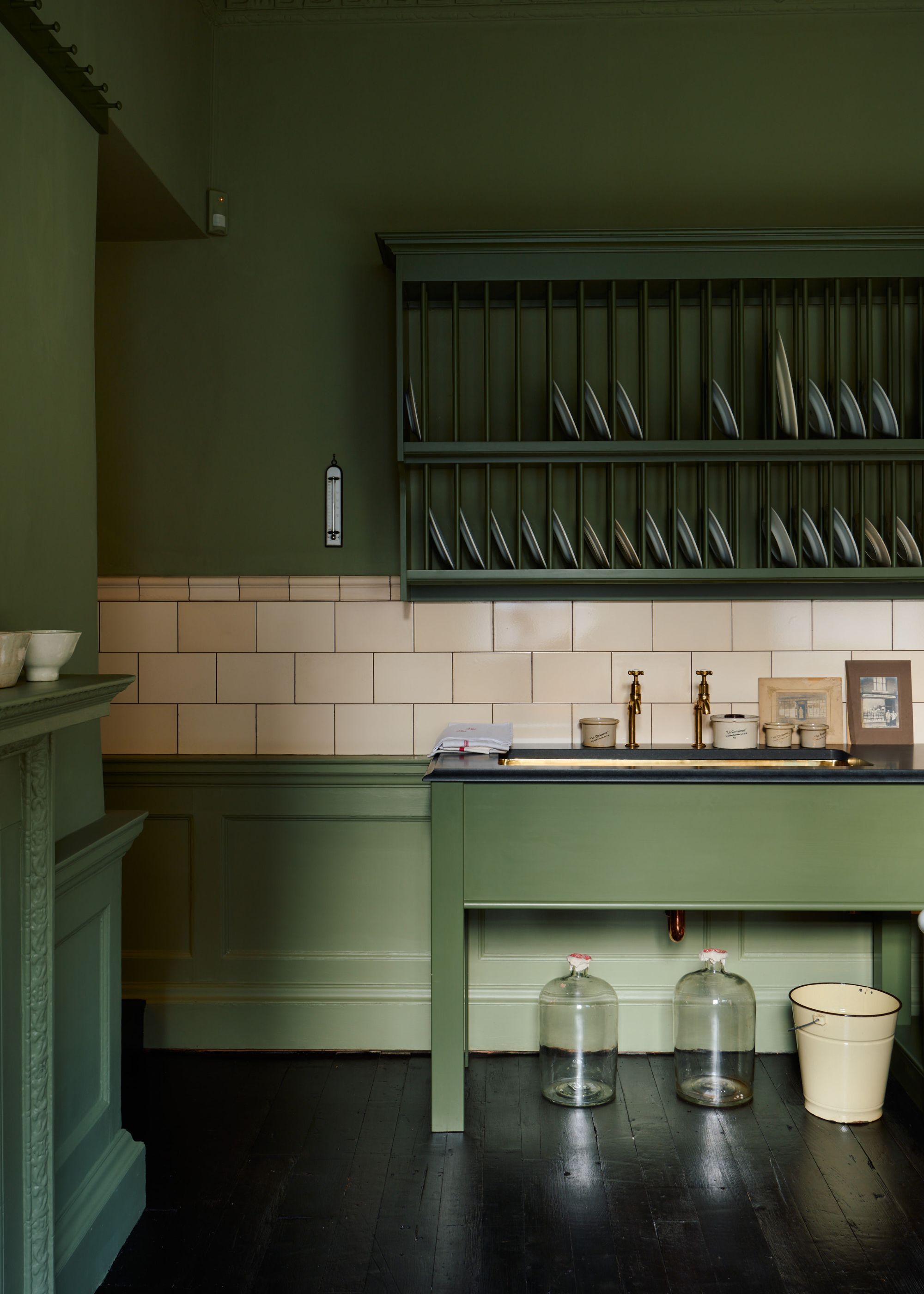 A deep green kitchen with a contrasting backsplash. In this kitchen is a two shelf plate rack with plates scattered around. Below the plate rack is a sink with two brass taps.