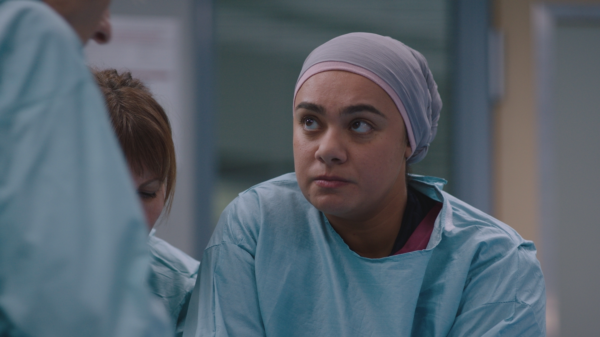 Rida in Surgical scrubs and a a light coloured surgical hijab looking intently at Russell.