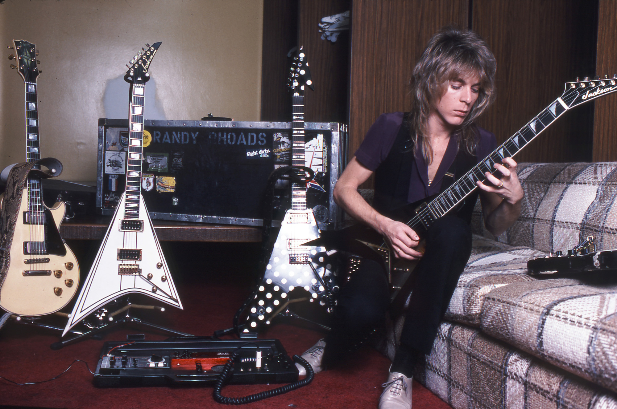 NOT FOR RE-USE!!!! A photo of Randy Rhoads practicing on the Diary of a Madman tour in early 1982. He's sitting alongside his 1974 Gibson Les Paul Custom, Jackson Concorde and Karl Sandoval Custom Polka Dot Flying V, all of which are on stands. A practice pedalboard, built for him by Zeus Audio Systems in Alhambra, California, sits in front of the guitars.