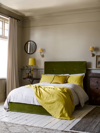 Neutral beige scheme, with a green velvet upholstered bed, white floorboards and bright citrus yellow lighting, pillows and throw.