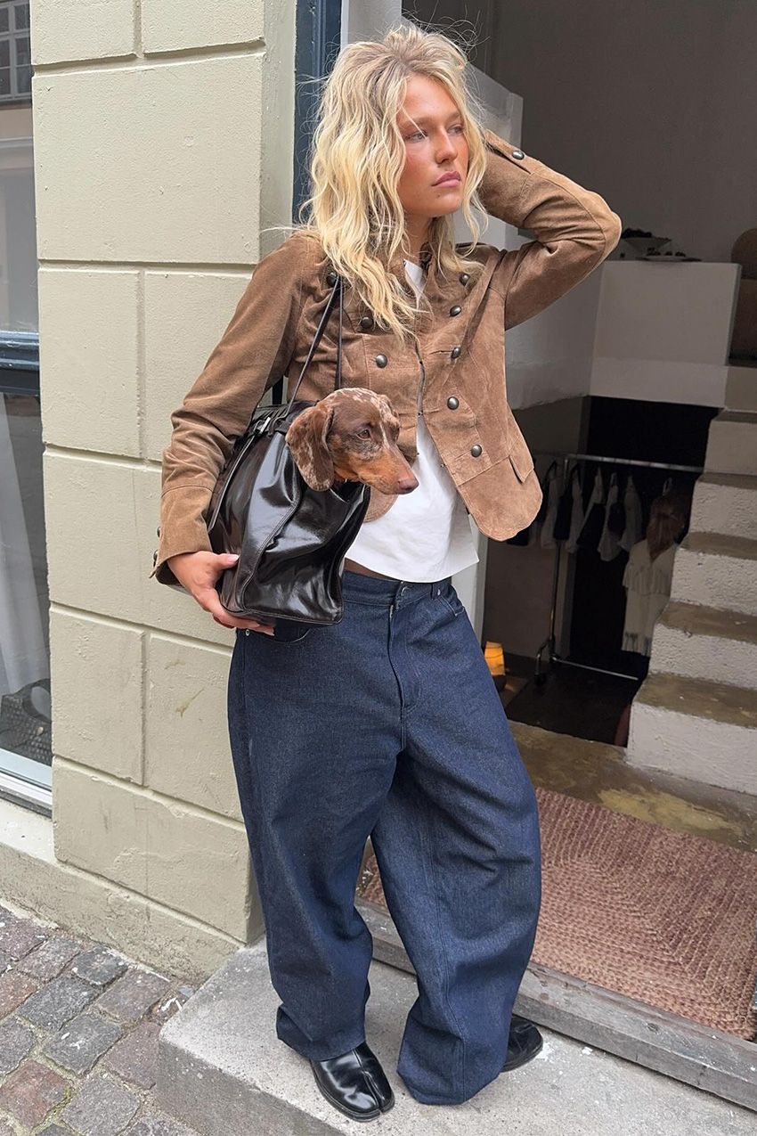 A street style photo of a woman standing in the doorway of a store wearing a suede military jacket over a white t-shirt, super baggy jeans, black loafers, and a black tote bag that has a puppy inside.