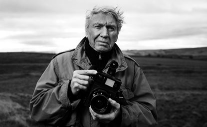A black an white photo of photo journalist Sir Don McCullin, a white male with short grey hair, wearing a large jacket and holding an old professional camera. 