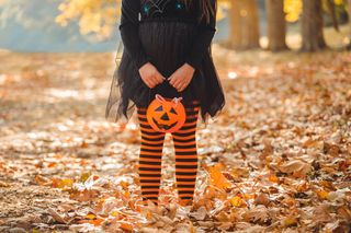 A young child wearing a witch costume with orange and black striped tights, standing amongst autumn leaves.