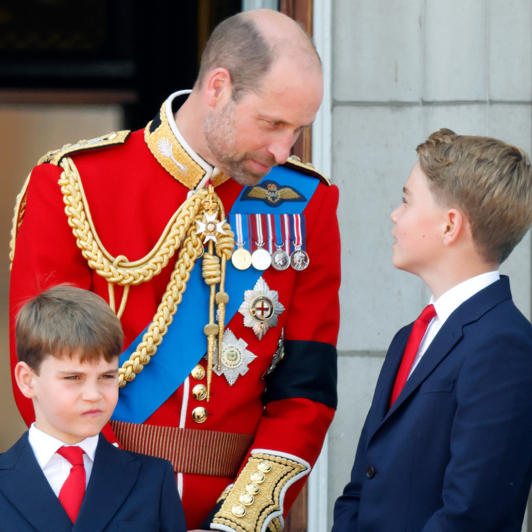 Prince William wearing a red uniform looking down at Prince George, who is smiling