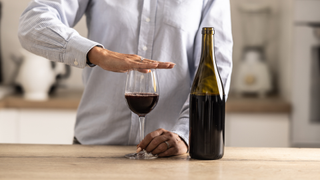 A bottle of half empty wine next to a full wine glass with someone&#039;s hand placed over the glass