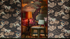 a design showcase featuring a lit table lamp with a red swirled base and orange lampshade on top of an abstract wooden side table with a circular top in an alcove decorated with ornate japanese style wallpaper and a green chair and the walls in front of the alcove are decorated with another ornate wallpaper with mythical creatures