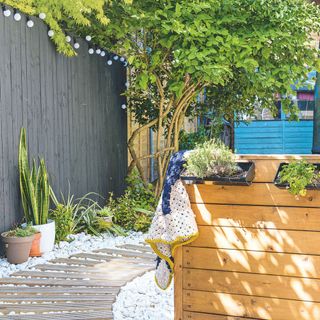 Garden area with white stones and a wooden walkway, surrounded by potted plants, trees, and a black painted fence