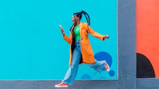 A woman is skipping gleefully against a bright background while looking at her phone