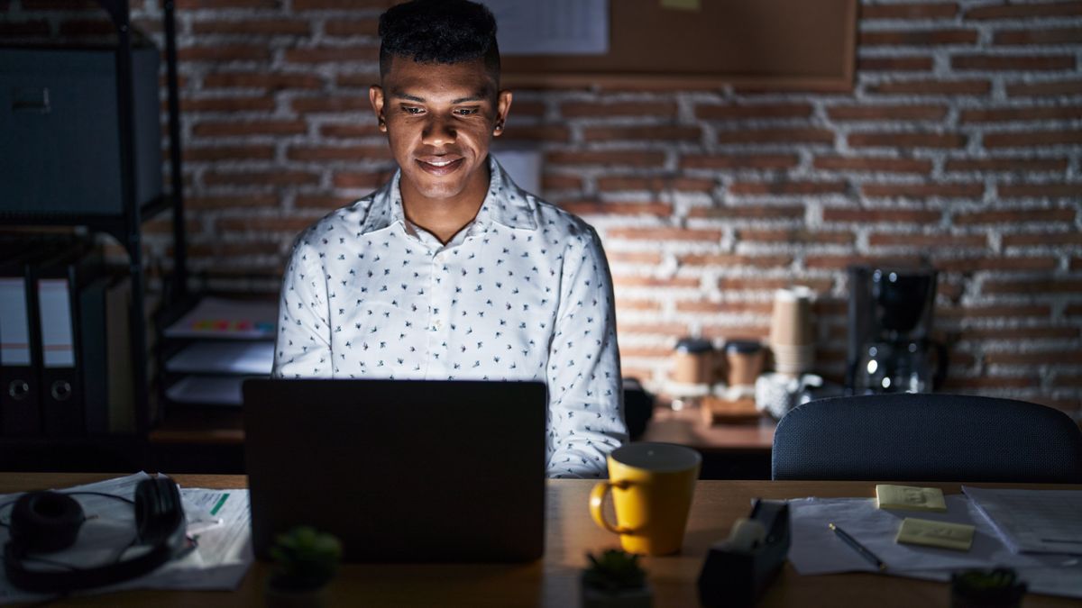 A man sat a table in a dark room, only list up by his laptop