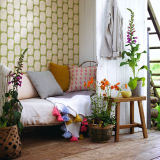 Wooden floored hallway with open white front door, daybed with cushions, flowers in pots and wallpapered wall