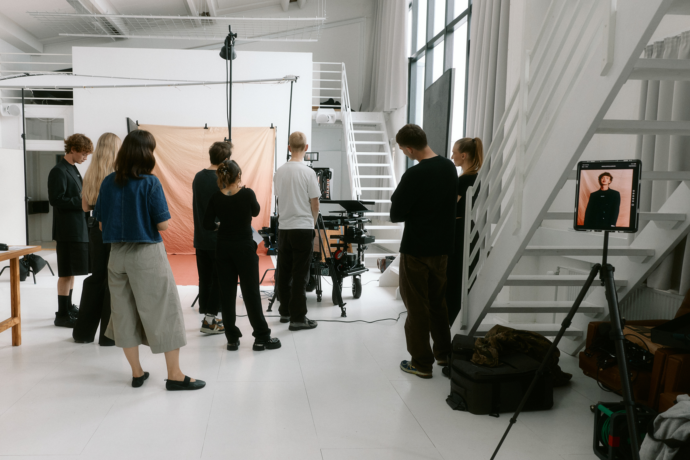 A group of people watches a photographer shoot a model against a peach backdrop in a bright, spacious studio with large windows