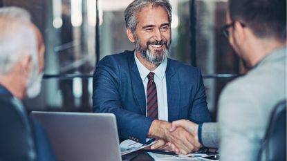 An older businessman smiles as he shakes hands with another man at a conference room table.