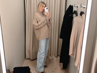 Natalie Cantell taking a dressing room selfie in a COS store in New York, wearing faded blue wide-leg jeans and an oversized beige sweater.