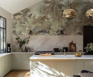 kitchen with marble splashback, tropical plant mural wallpaper above, pale green kitchen units and two gold pendant lights above kitchen island