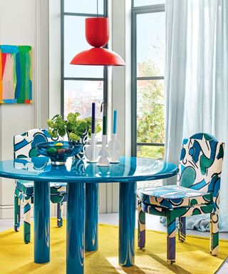 Colorful kitchen-diner, blue drapes, round blue dining table, upholstered patterned blue dining chairs, red pendant, colorful artwork in background