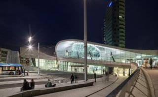 A night time view Arnhem Station with a curved double storey building with large glass walls, walkways and a tall building behind it.
