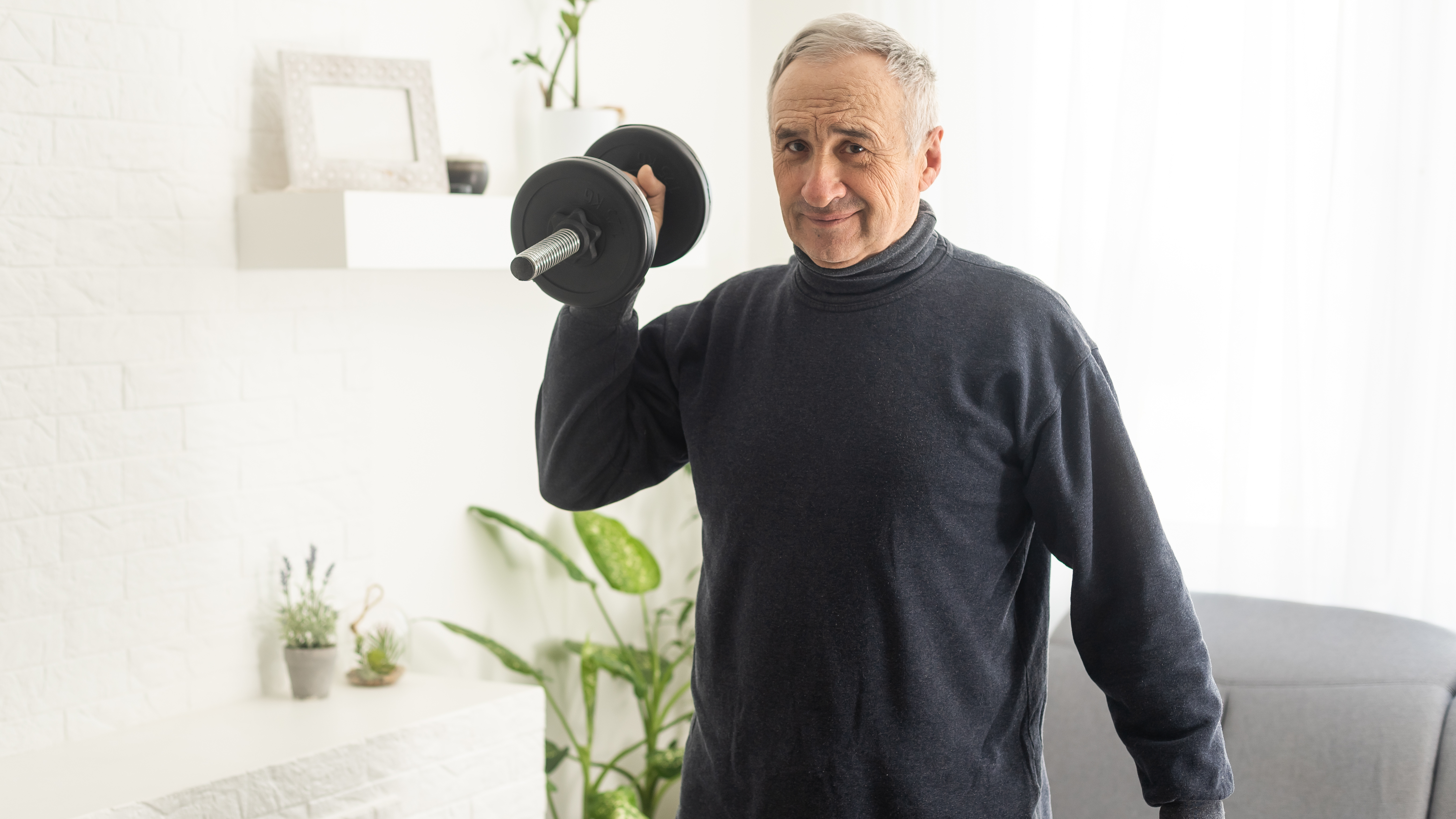 A man holds a dumbbell at his shoulder in a living room. Behind him we see a couch and plant.