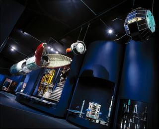 A museum gallery exhibits rockets, satellites and rocket engines against a dark blue backdrop