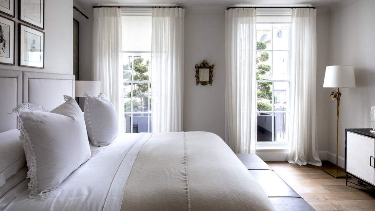 A white bedroom with a double bed and two tall windows with white crittal framing and floor to ceiling semi-transparent voile white curtains