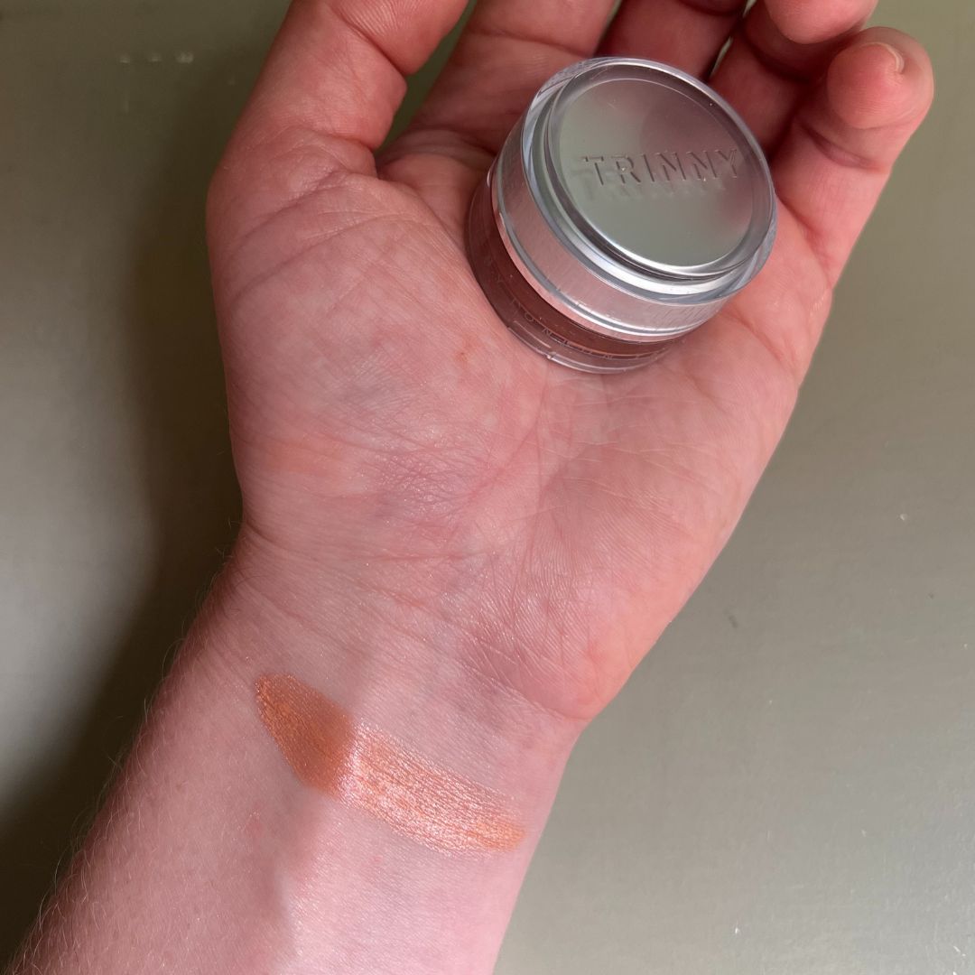 a hand on a green background holding Trinny London Eye2Eye cream eyeshadow with a swatch of the eyeshadow on the wrist 