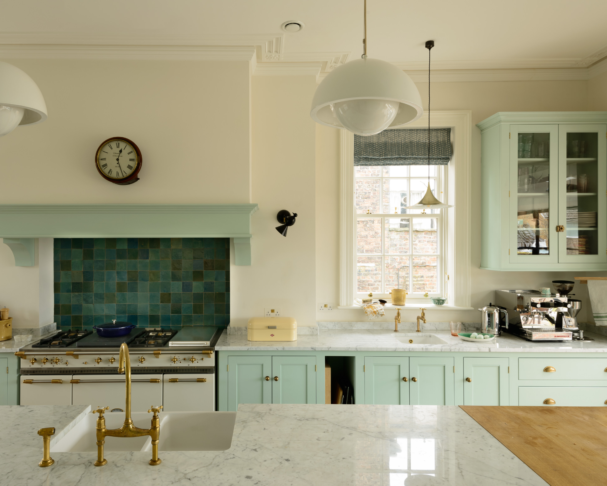 off white and pale green kitchen with tiled backsplash, marble countertops, sink in island, white stove, white pendant lights and glazed wall cabinet, coffee maker, black wall lights