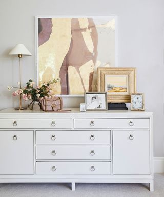 Corner of a bedroom that shows a white chest of drawers decorated with artwork, pictures, flowers, table lamp and ornaments, gray painted walls and carpet,